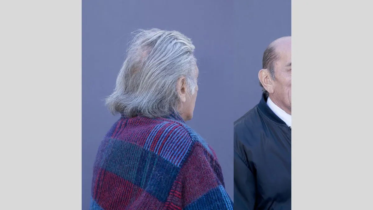  A man in his 70s, with styled grey hair faces away form the camera. He is standing in front of a grey blue background, wearing a red and blue tartan jumper and scarf.  Another man appears entering the right of the frame, he is bald and clean shaven and wears a white shirt and grey jacket.