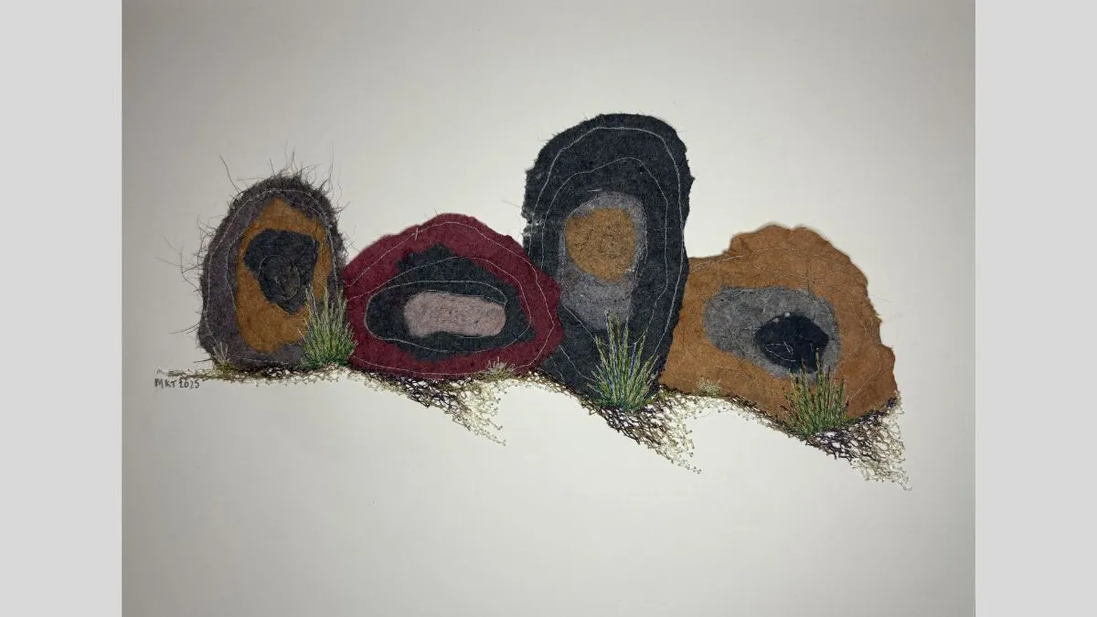 Horizontal textile artwork composed of five organic, stone-like forms in layered browns, greys, burgundy, and pink, stitched with visible thread and accented with tufts of green fibre, arranged against a light background.