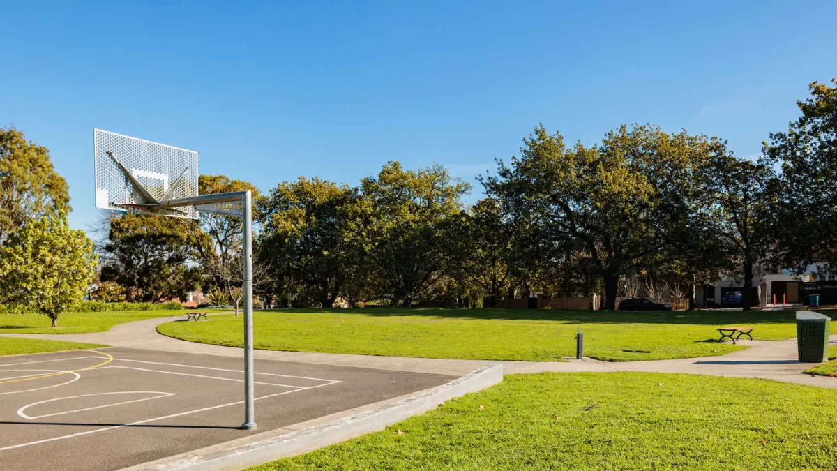 Part of a basketball court in a park with a concrete path and trees in the background