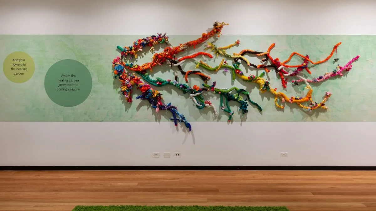 Wide wall-mounted artwork featuring twisting, branch-like forms made from brightly coloured wrapped fabrics and soft materials, set against a pale green wallpaper above a polished timber floor. Two green circles are positioned to the left of the work with text reading ‘Add your flowers to the healing garden’ and ‘watch the healing garden grow over the coming seasons. 