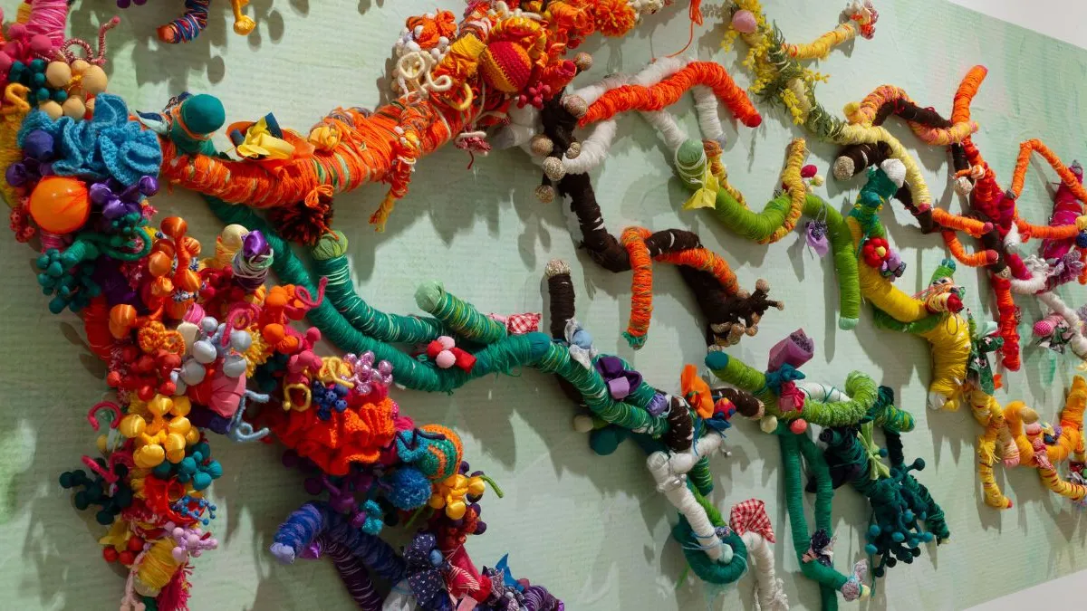 Close-up of a wall artwork showing intertwined, branch-like forms wrapped in vividly coloured yarns and fabrics, with clusters of soft, tactile materials, beads, and stitched elements layered densely against a pale green background.