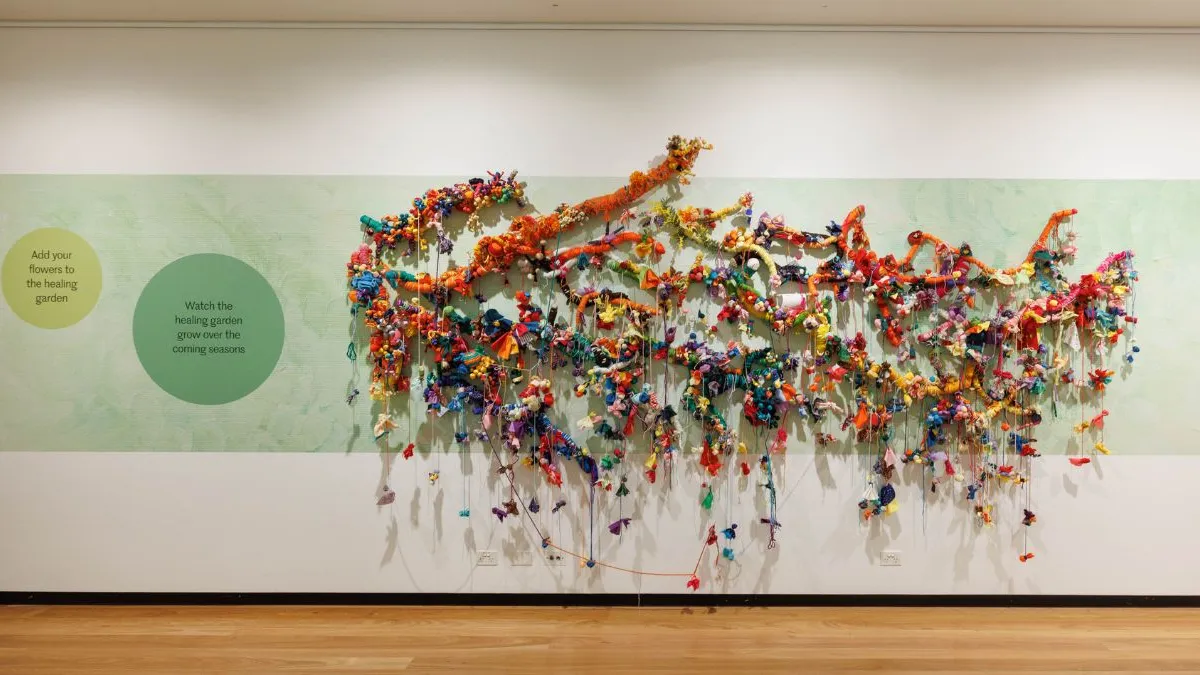 Wide gallery view of a large wall-mounted installation with branching, vine-like forms densely covered in colourful fabric, yarn, and hanging elements, extending across a pale green panel on white wall above a timber floor. Two green circles are positioned to the left of the work with text reading ‘Add your flowers to the healing garden’ and ‘watch the healing garden grow over the coming seasons.