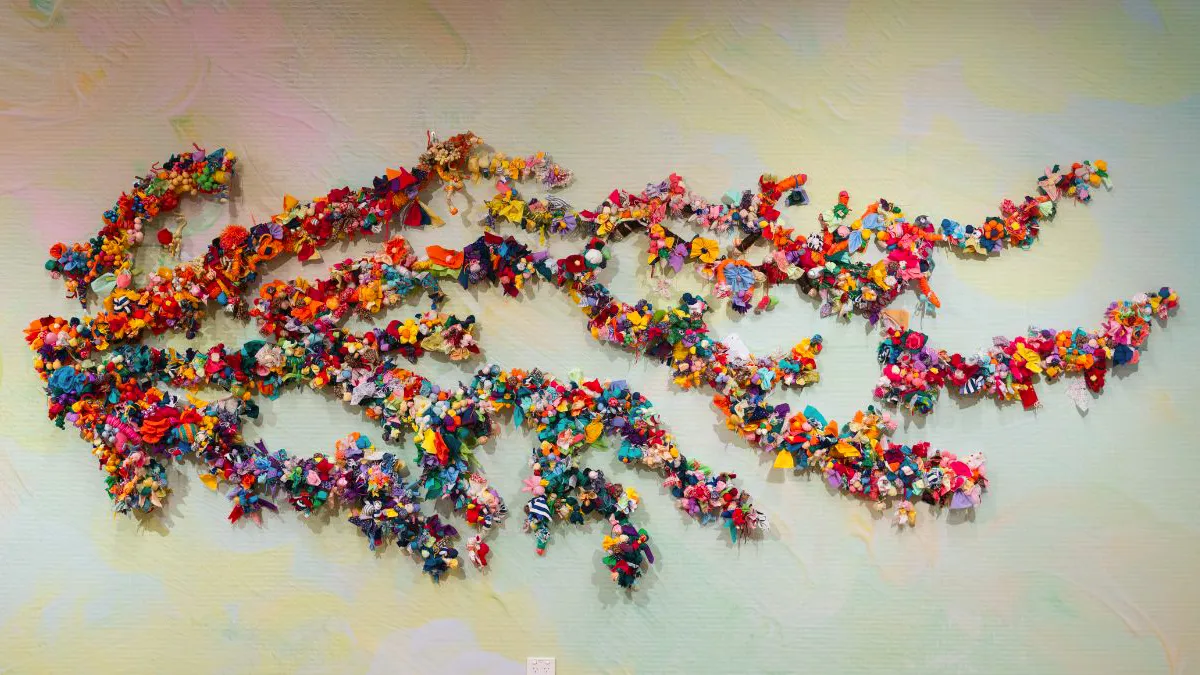 Wide view of a wall installation composed of branching forms densely covered in colourful fabric flowers and soft textile elements, arranged across a softly textured, pastel-toned background.