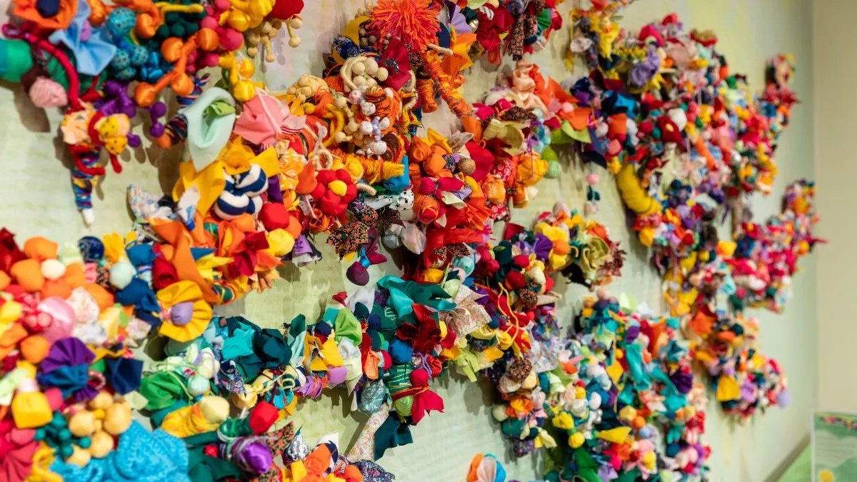 Close-up view of a wall installation showing densely layered, brightly coloured fabric flowers and soft textile forms clustered along branching structures against a pale green background.
