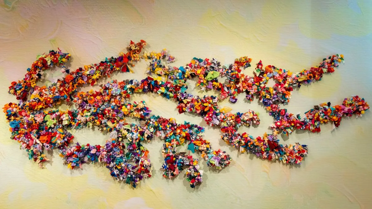 Wide view of a wall-mounted installation formed from branching shapes densely covered with colourful fabric flowers and soft textile elements, set against a softly blended pastel background.