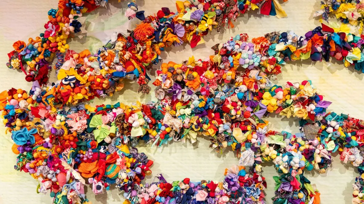 Close-up of branching forms densely layered with brightly coloured fabric flowers, knots, beads, and soft textile elements, creating a richly textured surface against a pale, softly patterned background.