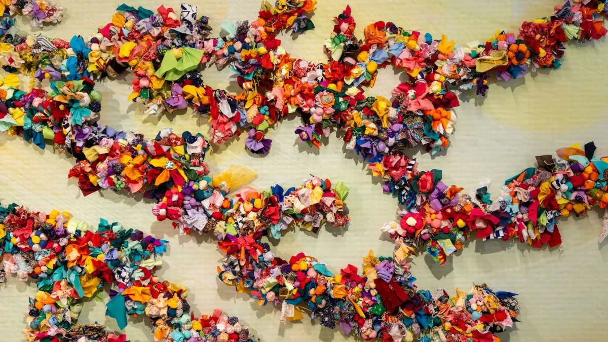 Close-up view of a wall installation showing branching lines densely covered with multicoloured fabric flowers, knots, and soft textile clusters arranged against a pale green, subtly textured background.