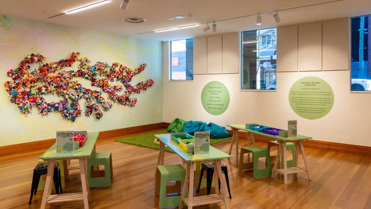 Wide gallery view showing a participatory installation with a large wall artwork of colourful fabric flowers arranged in branching forms, alongside green worktables with stools, material trays, beanbags, a rug, and wall text within a light-filled exhibition space.