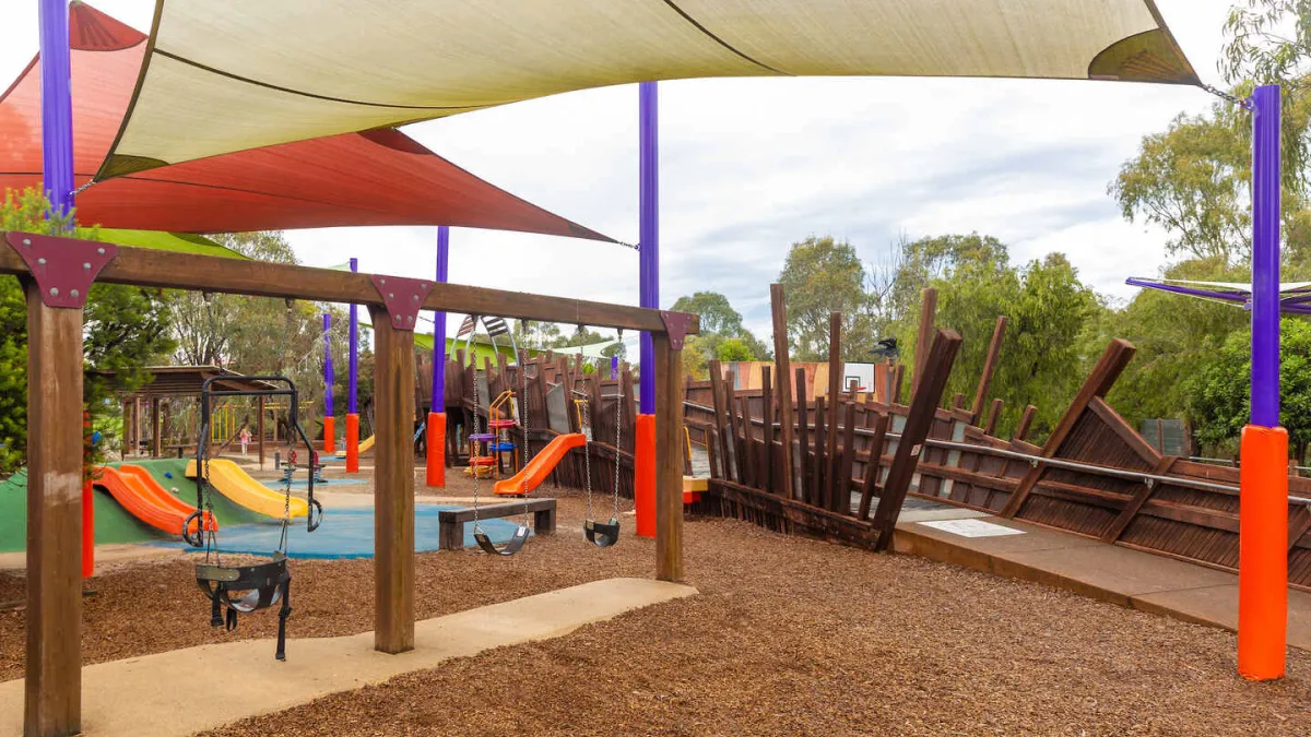 A playground with a wooden walkway, swings, slides under shade sails. 