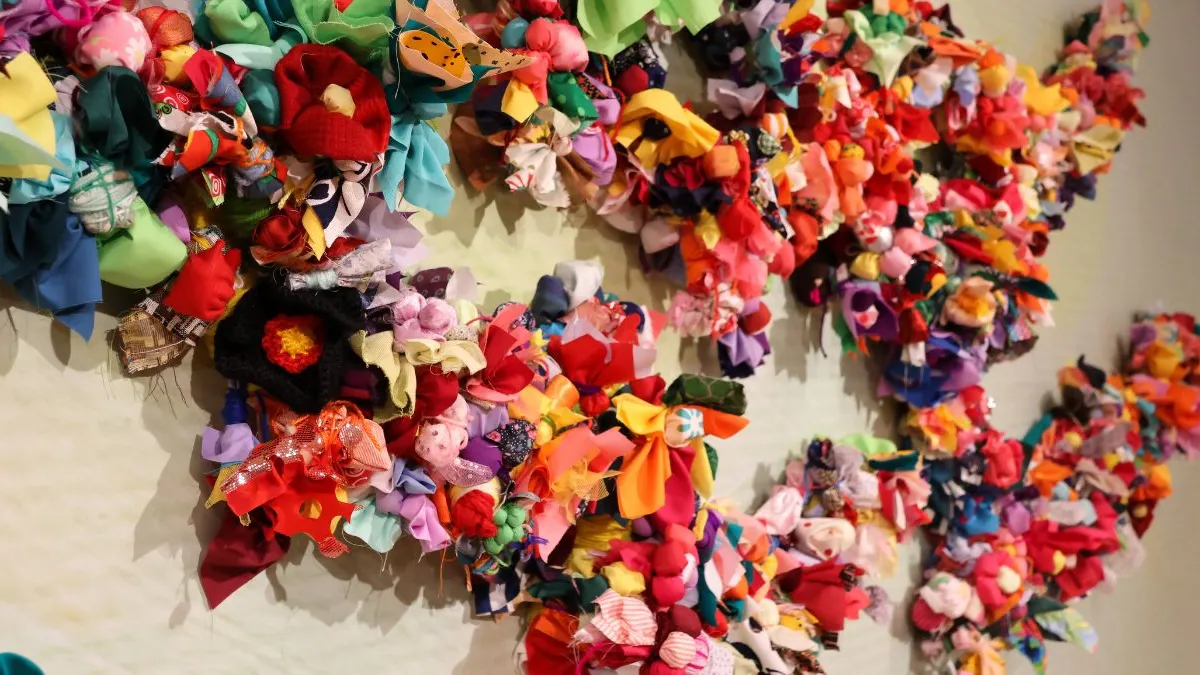 Close-up of a wall installation showing densely clustered, brightly coloured fabric flowers and soft textile forms arranged along branching lines against a pale, subtly textured background.