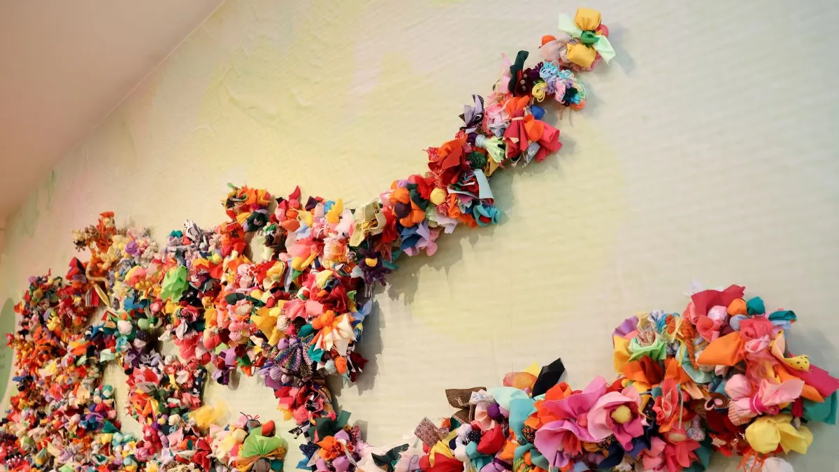 Angled close-up of a wall installation showing branching forms covered with densely layered, brightly coloured fabric flowers and soft textile elements against a pale, softly textured background.