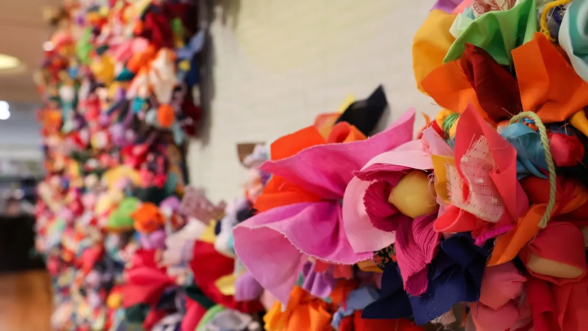 Shallow-focus close-up of a wall installation showing a dense cluster of brightly coloured fabric flowers and soft textile forms in the foreground, with additional clustered elements receding along the wall behind.