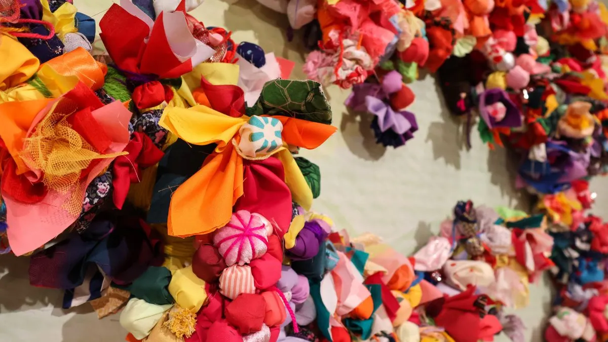 Close-up of a wall installation showing a densely layered cluster of brightly coloured fabric flowers, knots, and soft textile elements, with additional clustered forms visible in the background.