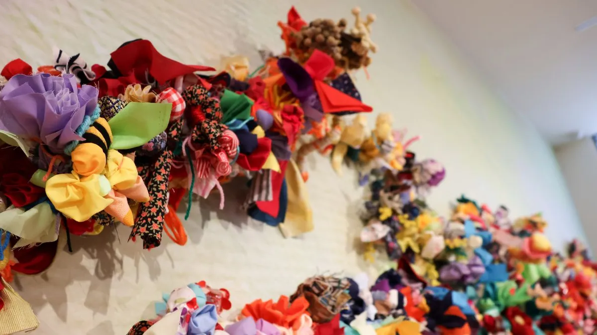 Angled close-up of a wall installation showing branching forms densely covered with colourful fabric flowers and soft textile clusters, receding along a pale, softly textured wall.