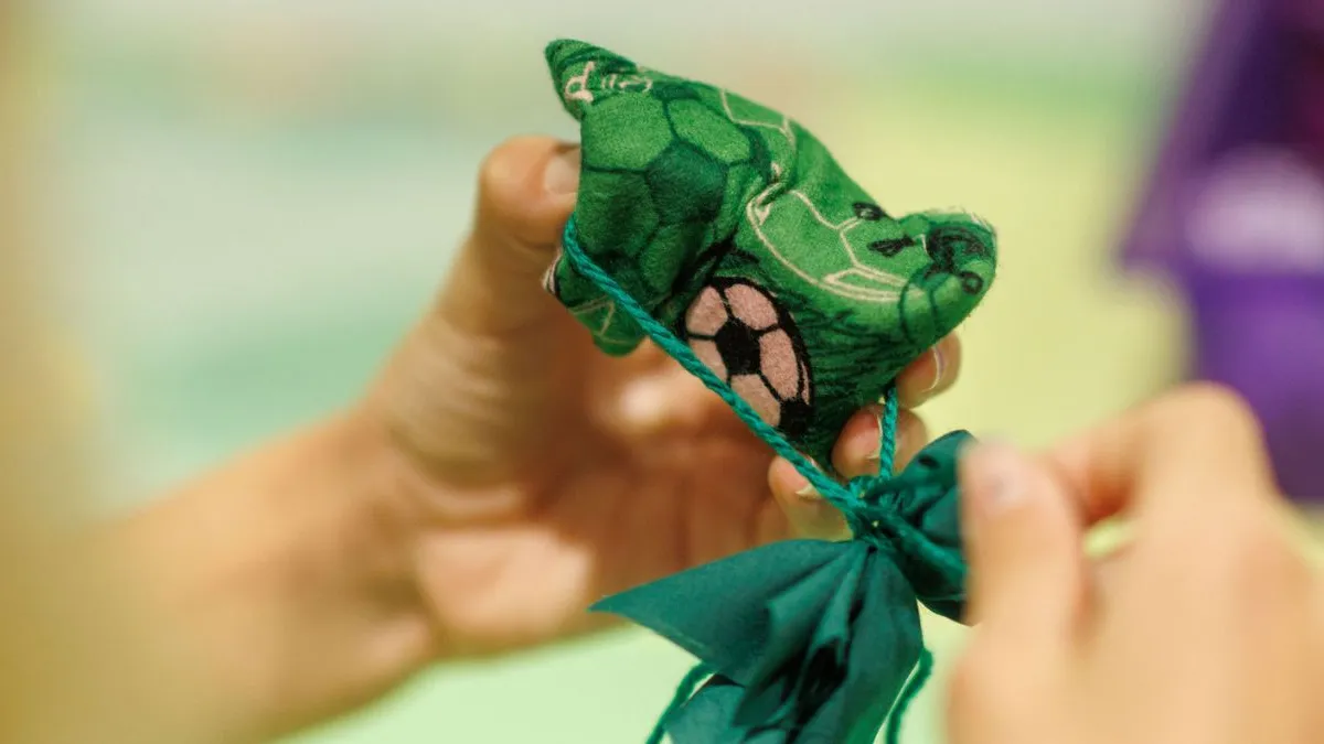 Close-up of hands tying a small, green fabric flower with string, showing soft textile folds and stitched details against a softly blurred background.