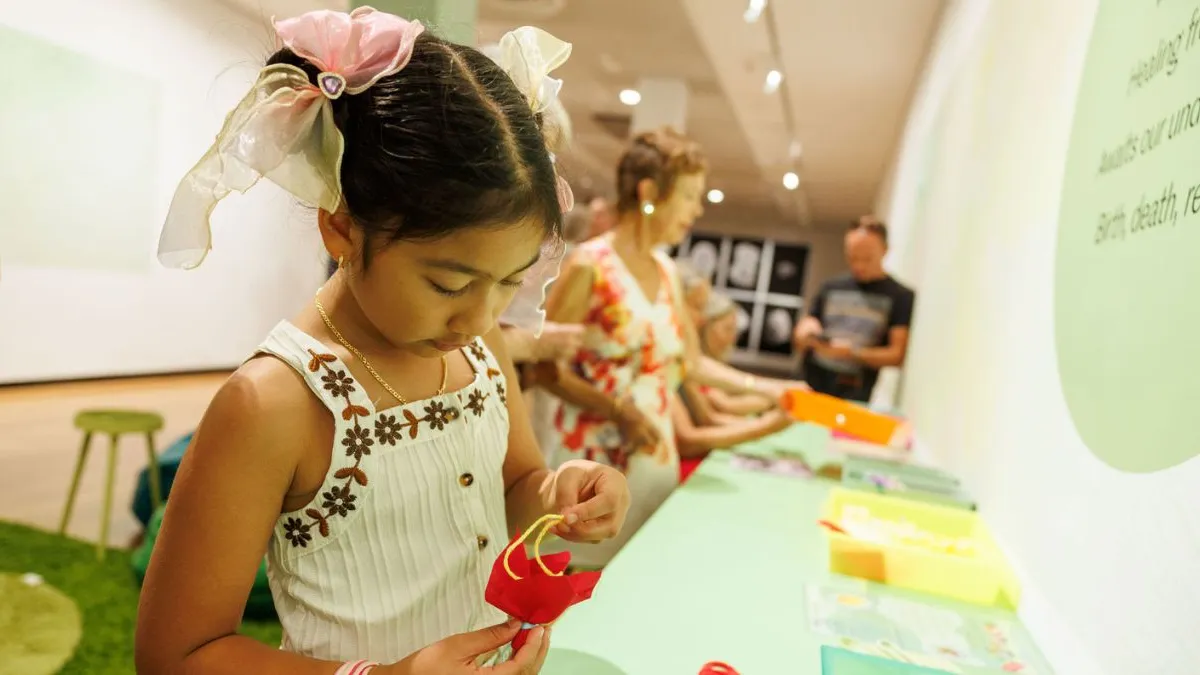 A young child concentrating while making a small fabric flower at a green worktable in a gallery, with other participants and materials visible in the background.