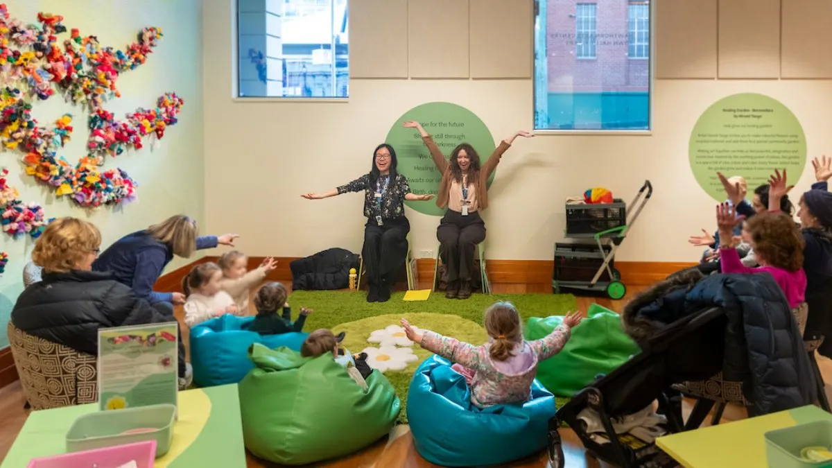 Group of young children seated in green beanbags facing two women. The women and children all have their hands in the air. The beanbags are arranged on a green rug with daisy pillows. To the left is a wall installation showing branching forms densely covered with colourful fabric.  