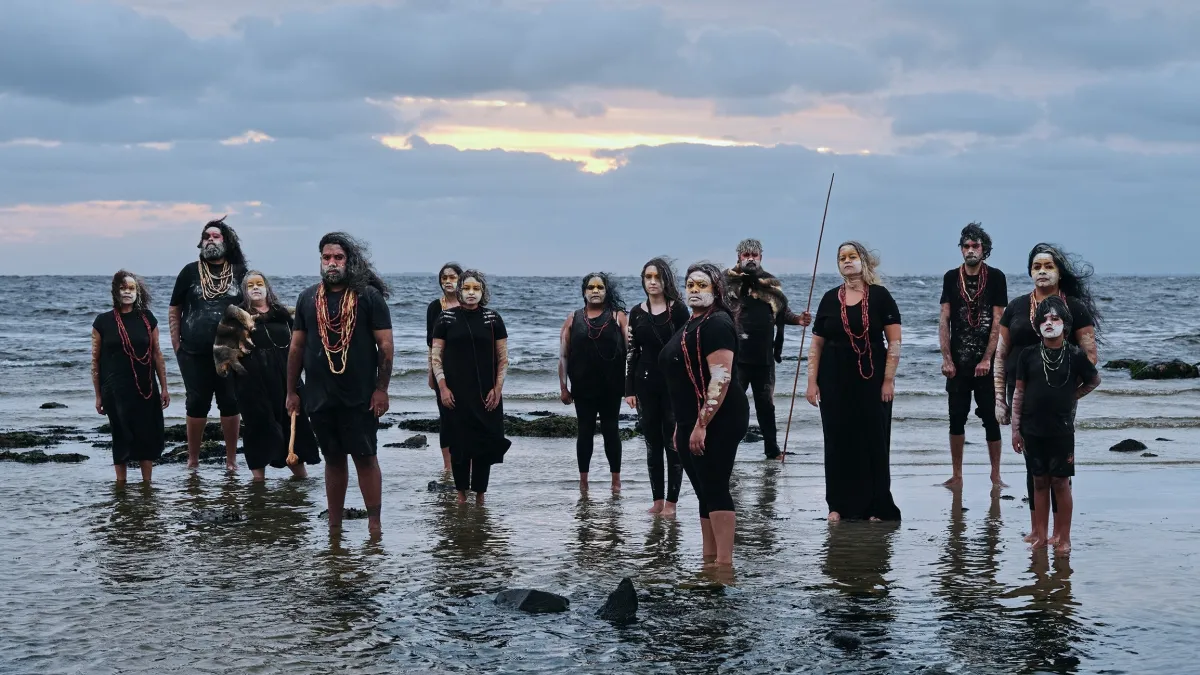 14 people in black clothing stand on the shore against the horizon. They wear river reed necklaces and their faces are covered in white and yellow pigment.