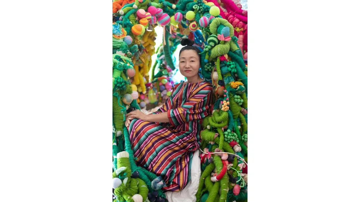 A woman sits inside a brightly coloured textile structure made of thick, braided yarn, forming a tunnel‑like space around them, wearing a striped, multicoloured, stripped garment.