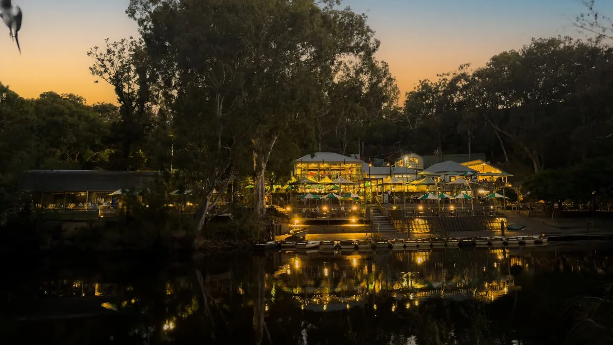 white umbrellas by a river at dusk. The building is surrounded by tall trees, and the sky is orange and blue. The lights of the building are reflected clearly in the still water below.