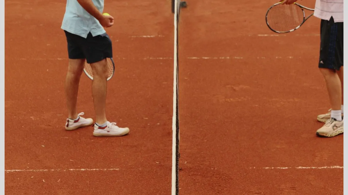 Photograph of two people standing on opposite sides of a tennis net on a red clay court, showing only lower legs, shoes, tennis rackets, and a ball, with the net running vertically through the centre of the photograph.