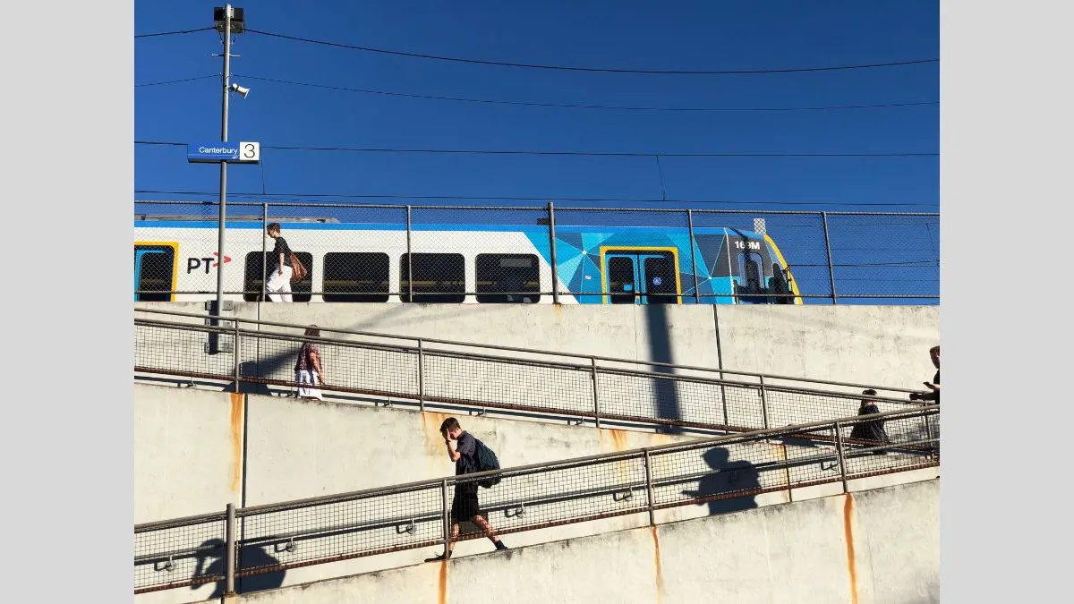 Photograph of a blue and silver train passing behind a fenced platform, with two concrete ramps diagonally stretching from one side of the photo to the other and then back across. There are igures moving on the platform and down the ramps, casting strong shadows under a clear blue sky. The sign in front of the train indicates the photo was taken at Cantubury station, platform 3.