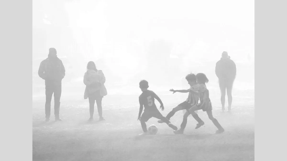 Black and white photograph of three children clustered together playing soccer through thick mist, with a soccer ball at their feet. There are two figures in the background to the left of the group and one figure standing to the right, all watching the game.