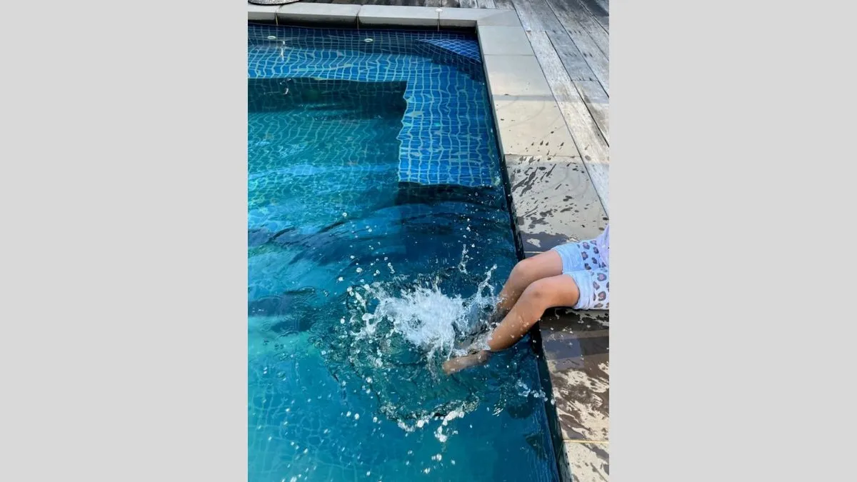 Photograph of a swimming pool with blue tiles and grey paving stones along the right-hand edge. Two legs belonging to a child in blue board shorts are seated on the edge of the pool. There is a large splash where the child's feet meet the water, and rippling water surrounds.