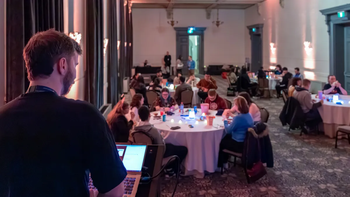 Looking from behind the quizmaster into a room with 4 large round tables. Each table has about 6 people working together.
