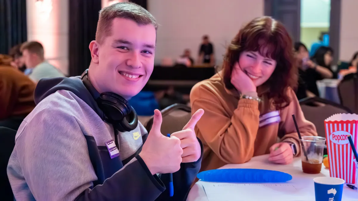 Two people sitting at a table with drinks and popcorn smile at us. The person closest has both their tumbs up and has headphones around their neck.