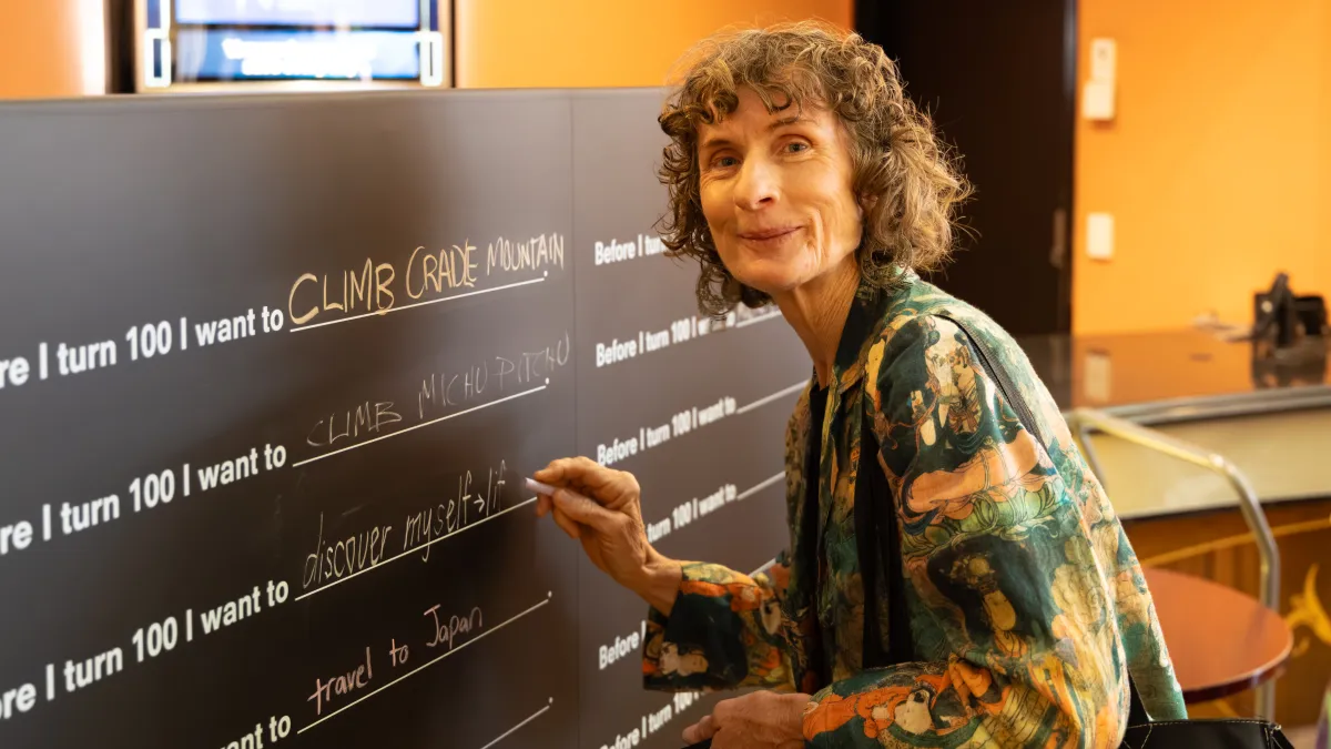 Person with short, curly hair looks at us with a smile while writing with chalk in response to the prompt "before I turn 100 I want to..."