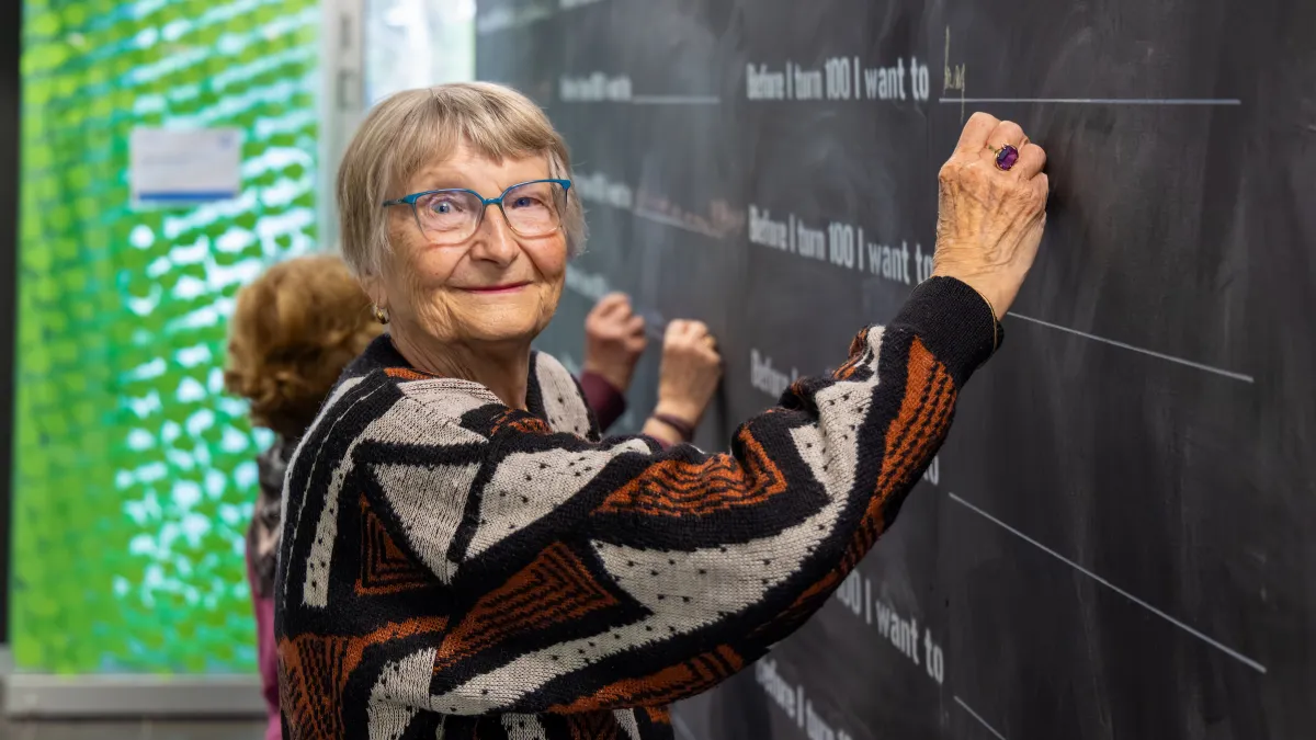 Woman wearing a brown and cream geometric patterned jumper writing on a wall with chalk what she wants to do before she turns 100.