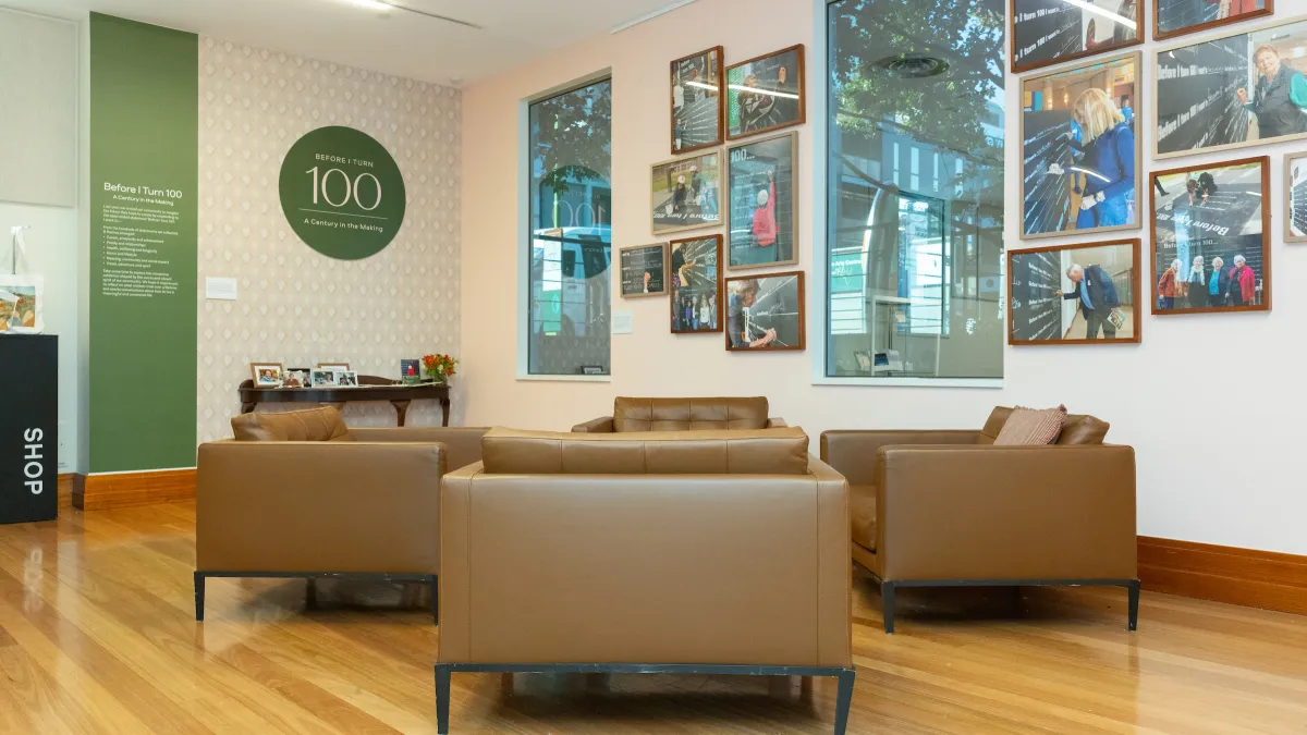 4 padded chairs are placed facing each other. in the wall behind are two tall windows and framed images of people writing what they want to do before they turn 100.