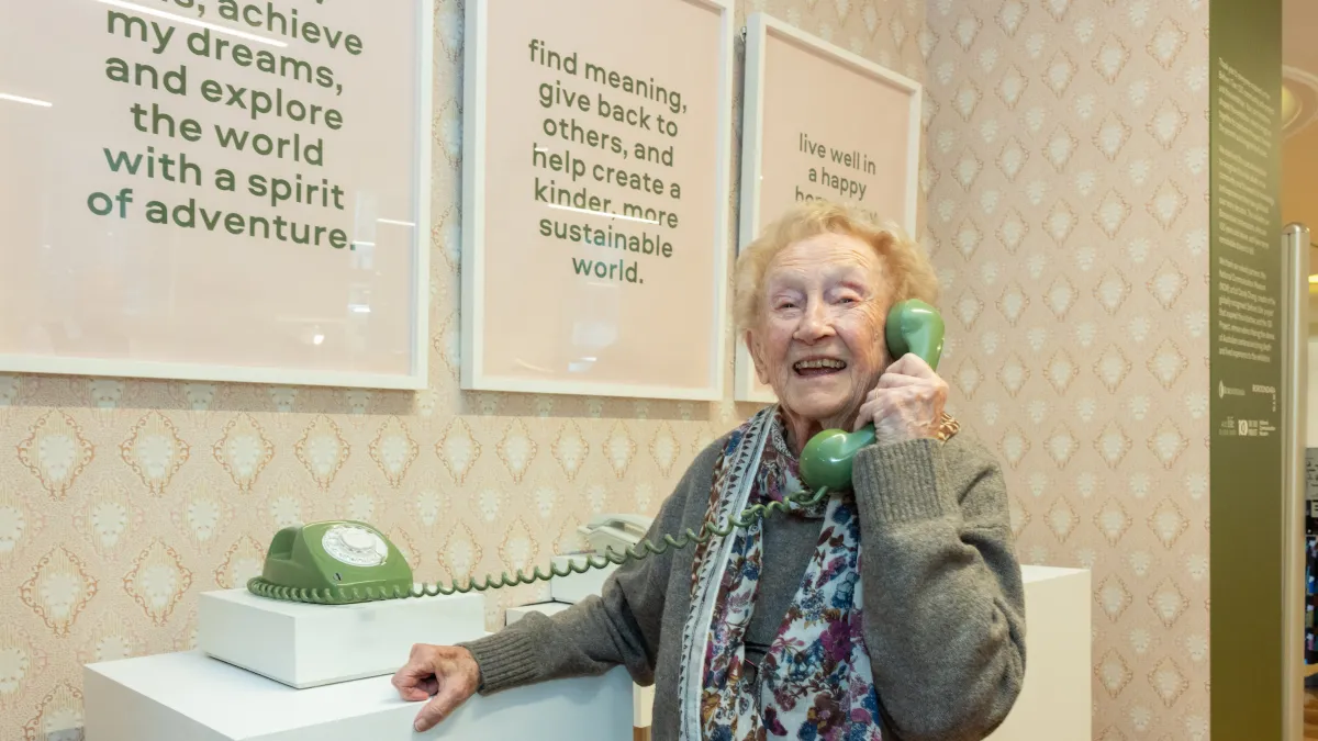 Woman holing a corded phone handpiece to her ear. the phone is green, and placed against a cream patterned wallpaper. On the wall are 3 frames with text.