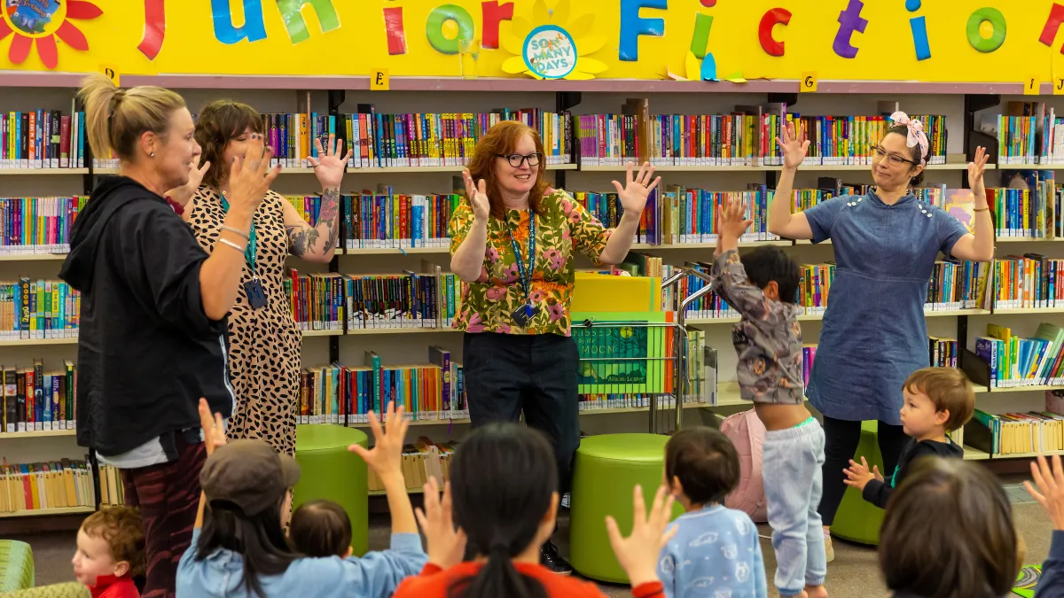 4 adults stand at the front of a group of children, all signing applause. In the background are shelves of books with the sign 'junior fiction' above them.