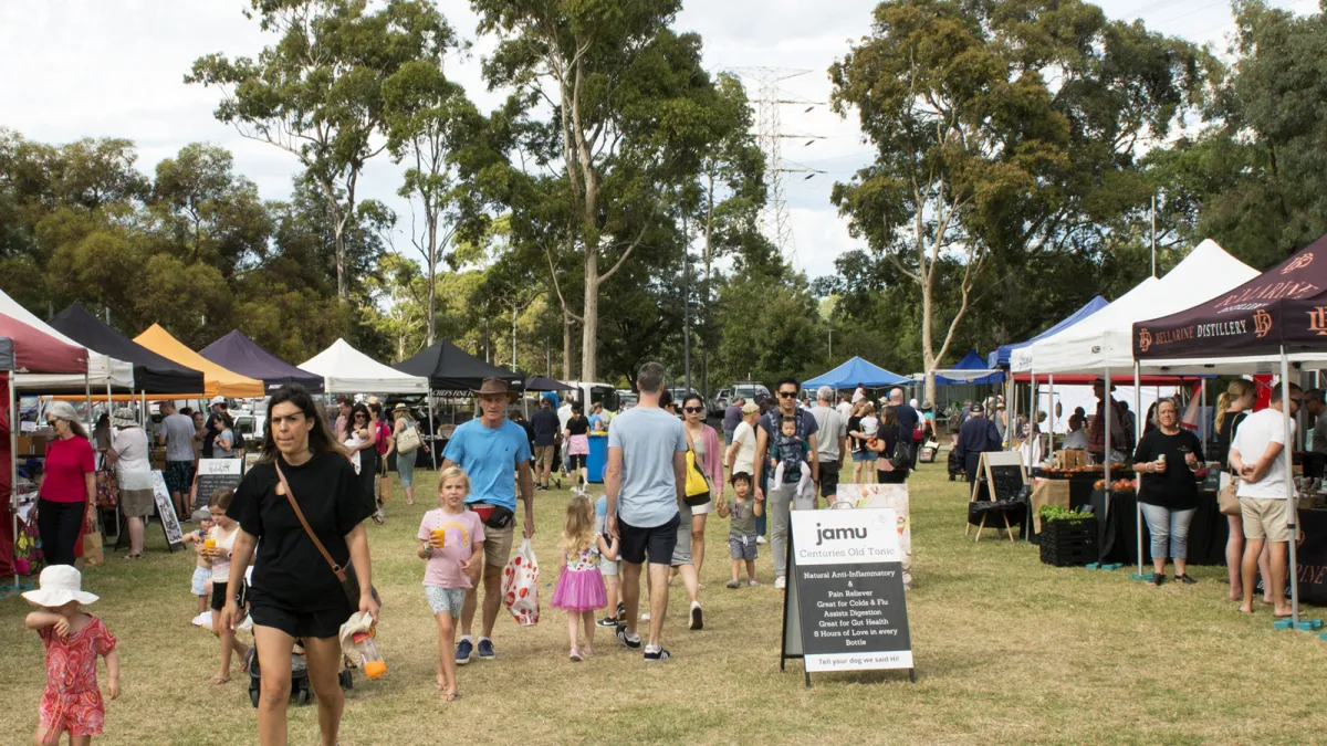market stalls line both sides. Families walk between stalls looking at and buying products. In the background are the tall gum trees of Patterson Reserve.