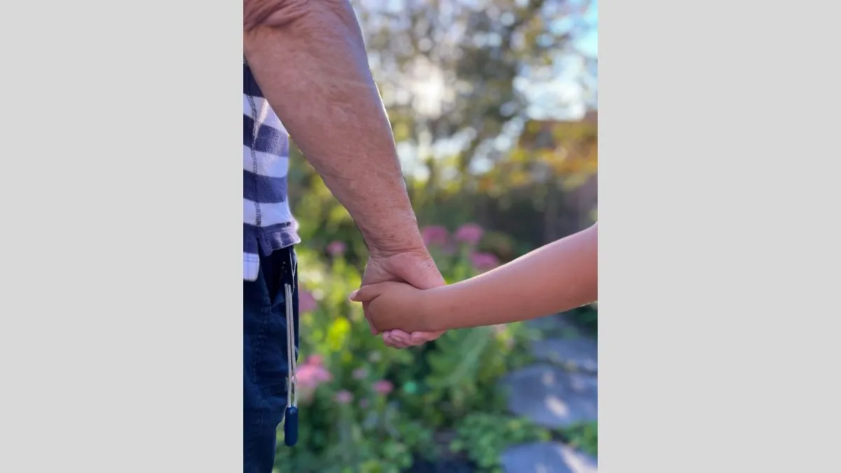 A close-up photograph of two arms stretched out holding hands. One arm belongs to an adult, and the other belongs to a child. The background is blurred and features green garden plants, stepping stones, trees and the sun.