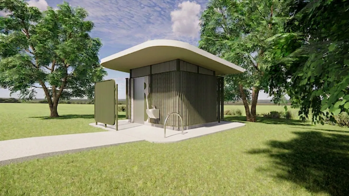 Front view of a green and cream toilet block. Large trees surround the block. There's a bike rack out the front.