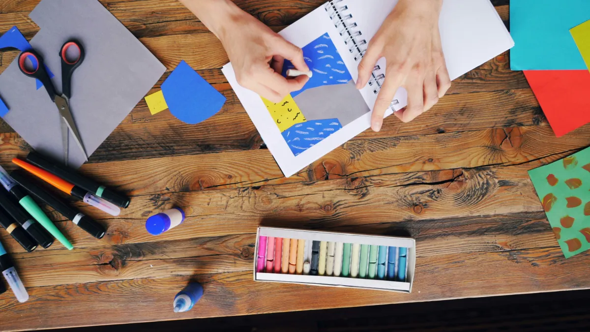A selection of pencils, crayons, scissors and paper sit on a wooden table with 2 hands visible, 1 holding a crayon