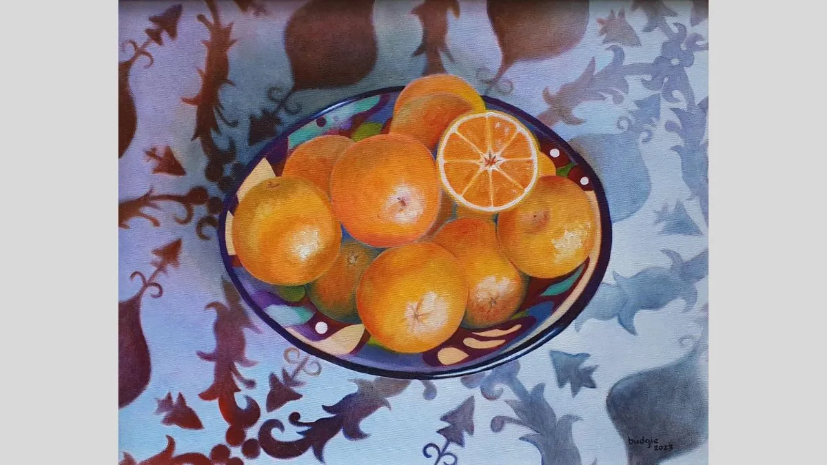 A still-life painting of a bowl filled with oranges. The bowl sits on a patterned cloth with floral and leaf shapes in muted red and grey tones. One orange is cut in half, showing pale segments. The bowl has a multicoloured rim with decorative shapes.