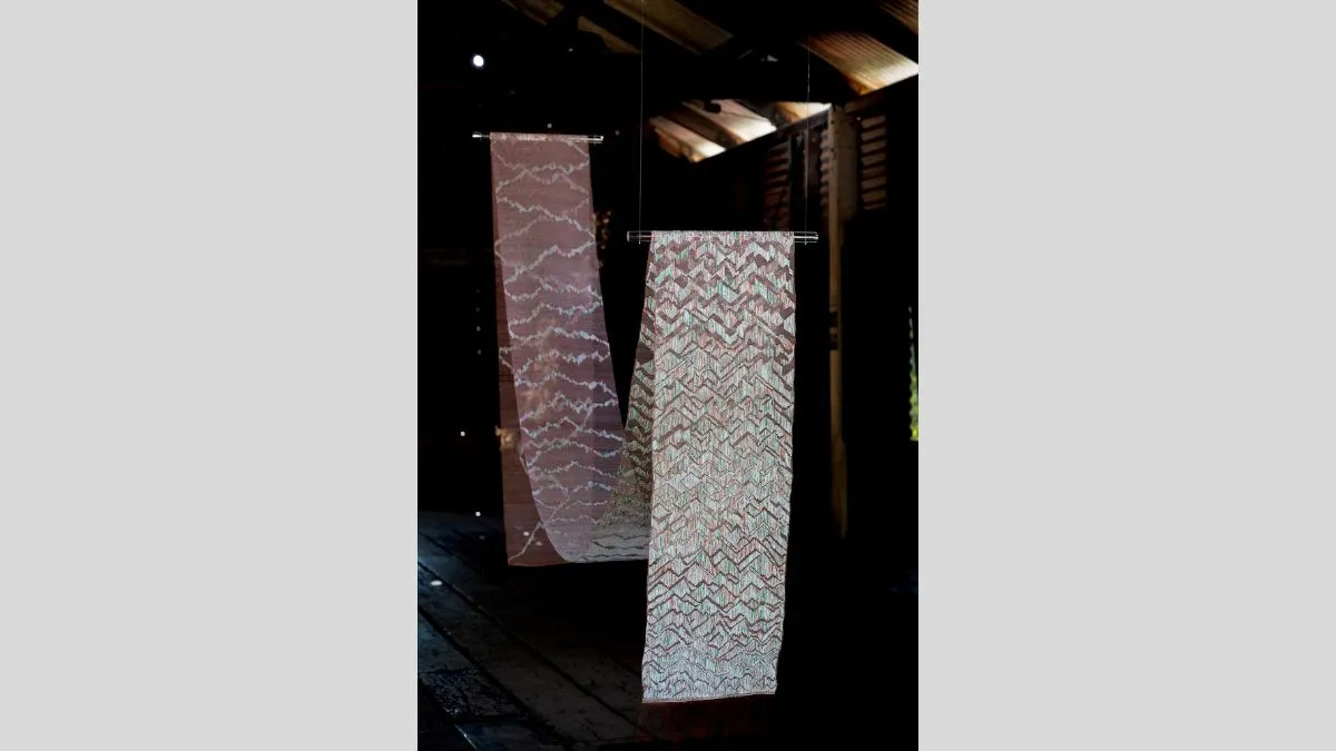 Two narrow woven panels hang vertically from clear rods in a dim, rustic interior with timber beams and floorboards. The panels feature repeating zigzag patterns in muted earthy tones, pale green and red. Light filters across their textured surfaces.