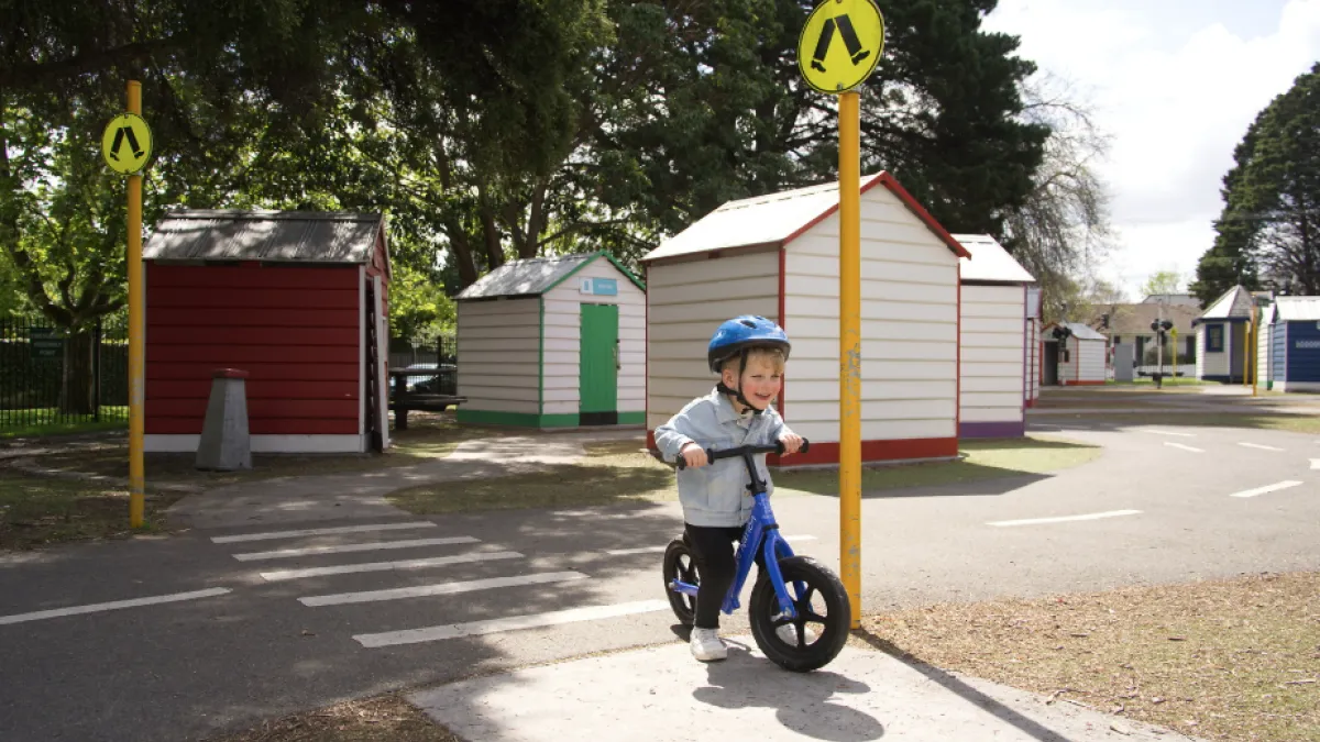 Child wearing a denim jacket and blue helmet rides a balance bike across a zebra crossing. In the background are small buildings.