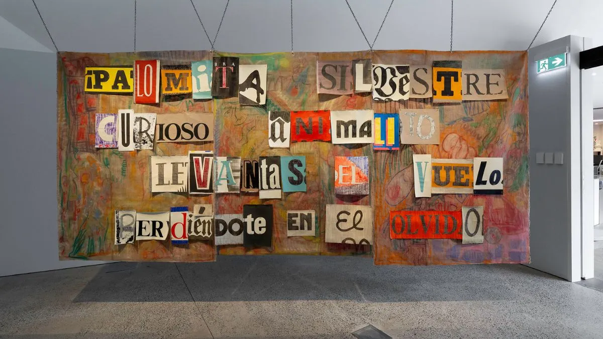 A large mixed media artwork installed in a gallery space. The surface is divided into canvas panels with doodles in orange, red, green, and brown. Across the panels, cut out letters form the Spanish text  “!Palomita silbestre curioso animalito levantas el vuelo perdiéndote en el olvido”. 