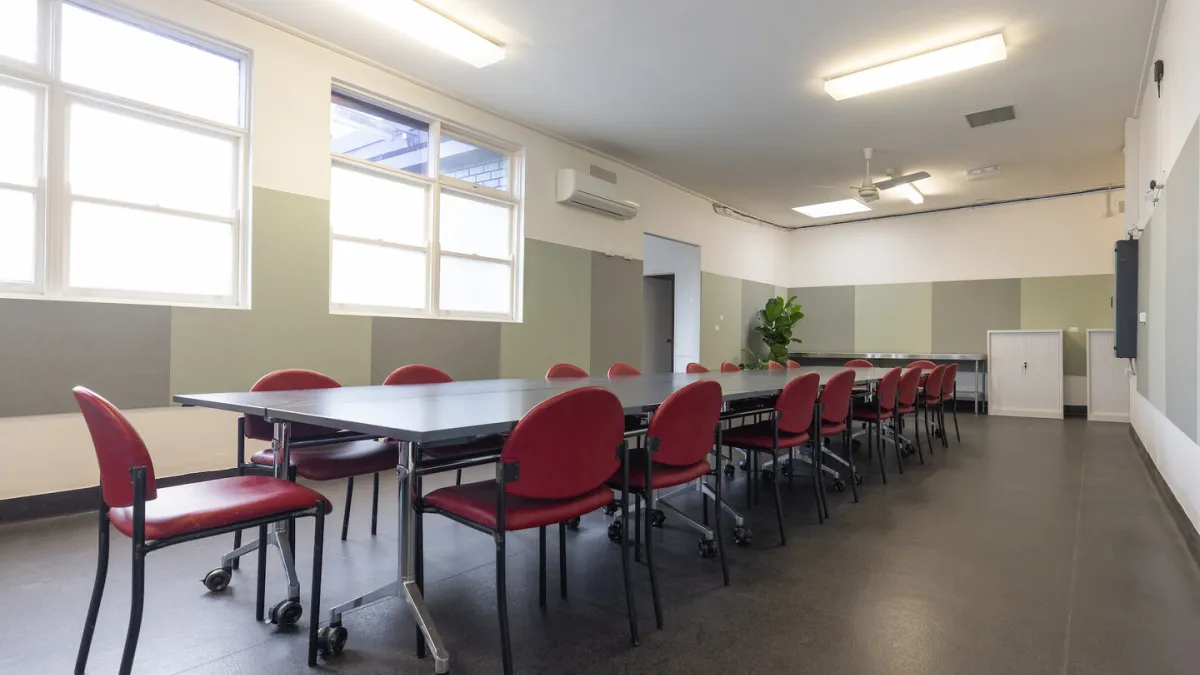 A meeting room with a long table surounded by red chairs. The room has a split system and a ceiling fan. 