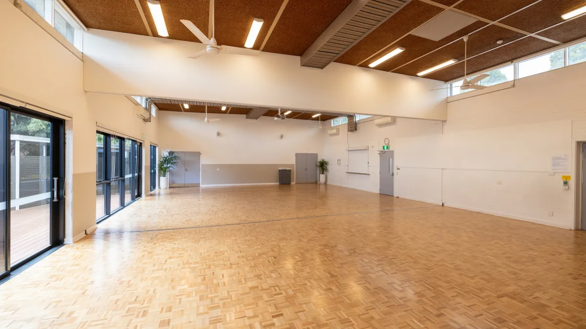 A large hall with wooden floors. There are windows on the left looking out to a decked area. 