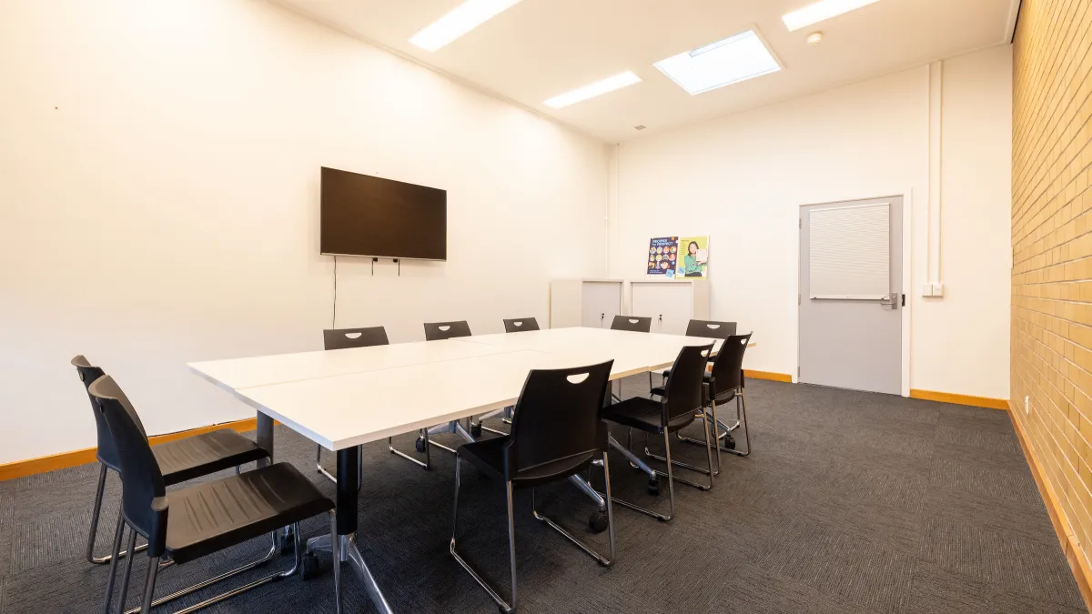 A meeting room with a table surrounded by chairs. It has a TV. 