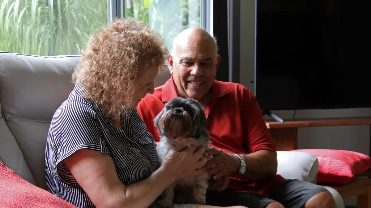 Two people sit on a couch smiling and patting a small black and white dog.