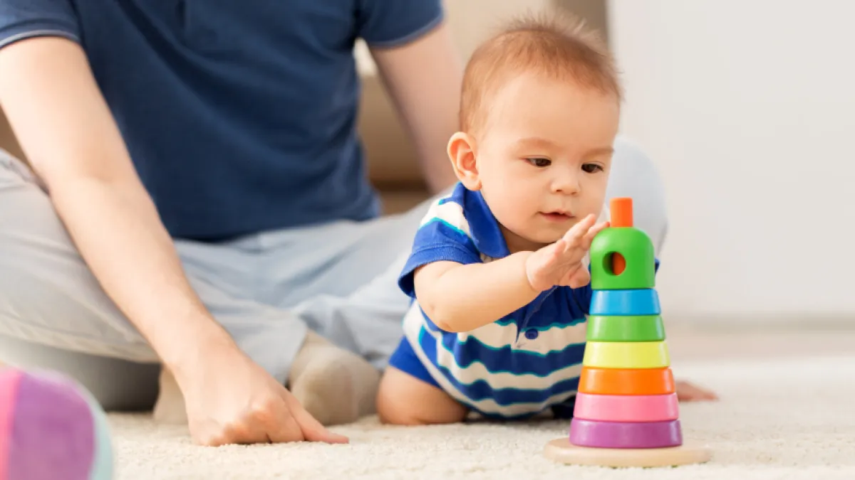 A baby plays with a colourful stacking toy while an adult sits nearby