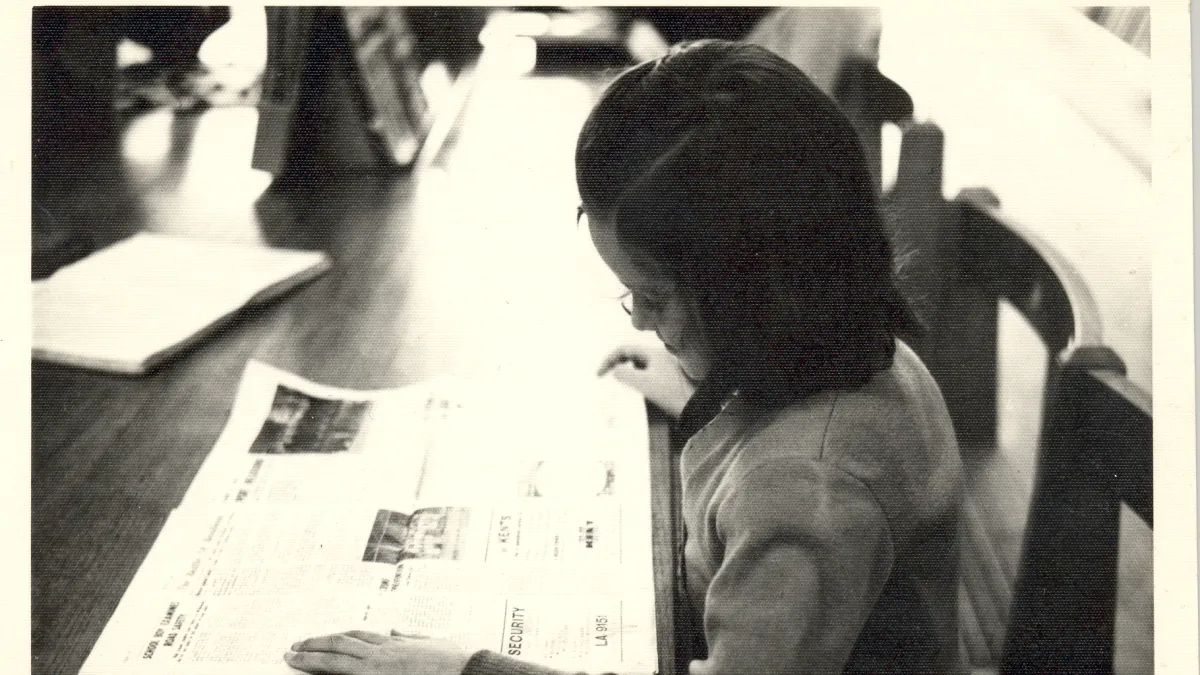 A black and white image shows a young child sitting at a wooden desk reading a newspaper