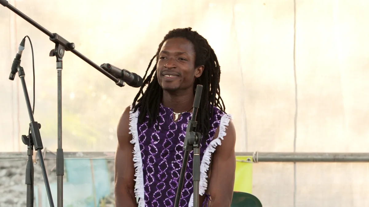 A man with long dark hair sits in front of a microphone. He is wearing a purple sleeveless top with white edging.